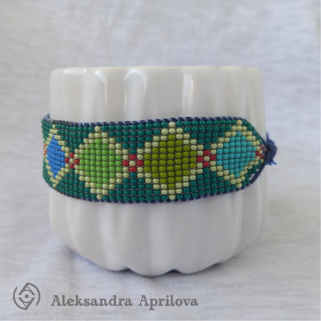 Дамска гривна от мъниста „Ариел“ Александра Априлова. Ръчно изработена, свежа, дамска гривна с геометрични фигури (ромбове) в студената цветова гама и с леко червено присъствие, подходяща за летния сезон. Авторски дизайн. Дизайнът на гривната е вдъхновен от любимата на всички ни анимация „Малката русалка Ариел“ като той изобразява нюансите на морските дълбини и красотата на изящните митични същества.