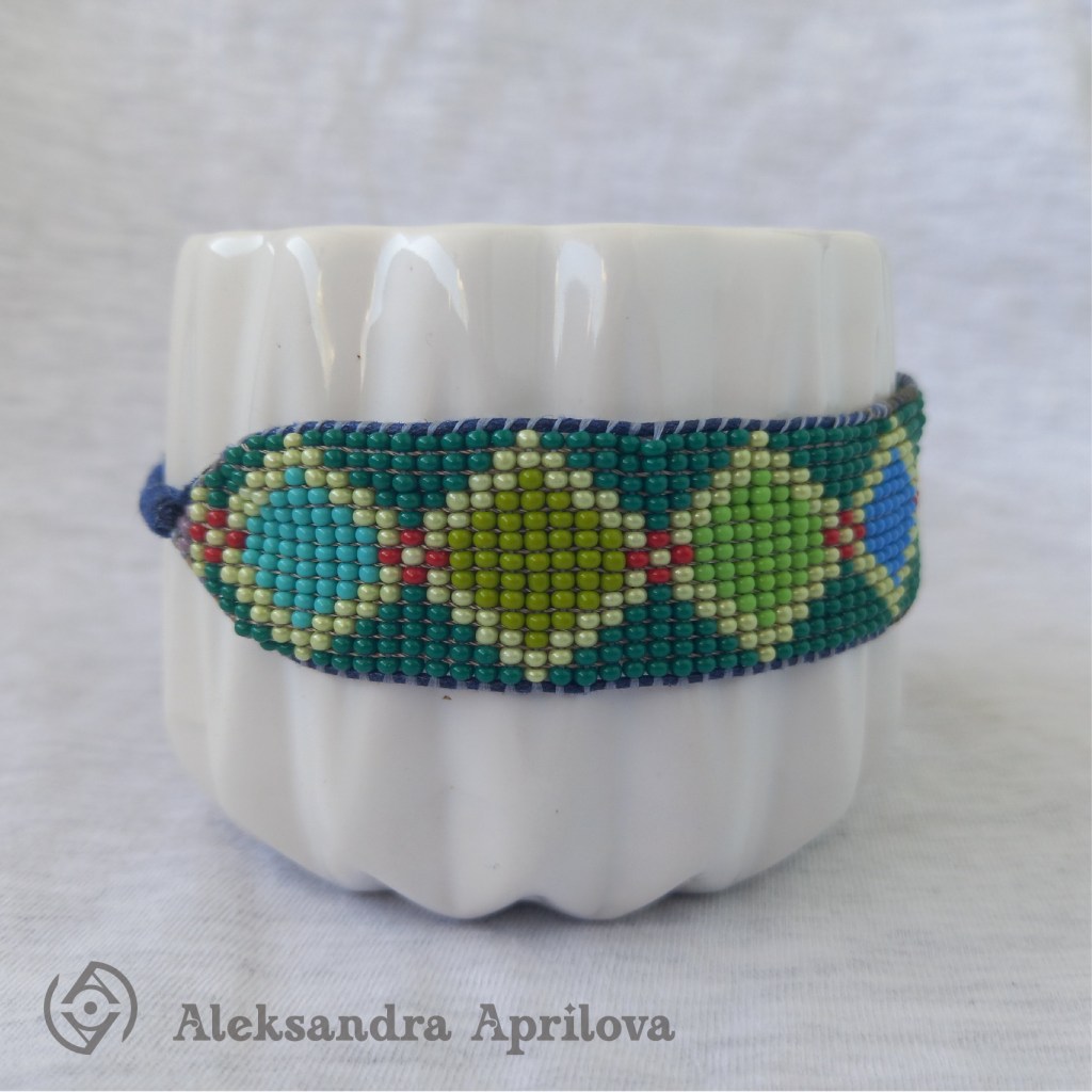 Дамска гривна от мъниста „Ариел“ Александра Априлова. Ръчно изработена, свежа, дамска гривна с геометрични фигури (ромбове) в студената цветова гама и с леко червено присъствие, подходяща за летния сезон. Авторски дизайн. Дизайнът на гривната е вдъхновен от любимата на всички ни анимация „Малката русалка Ариел“ като той изобразява нюансите на морските дълбини и красотата на изящните митични същества.